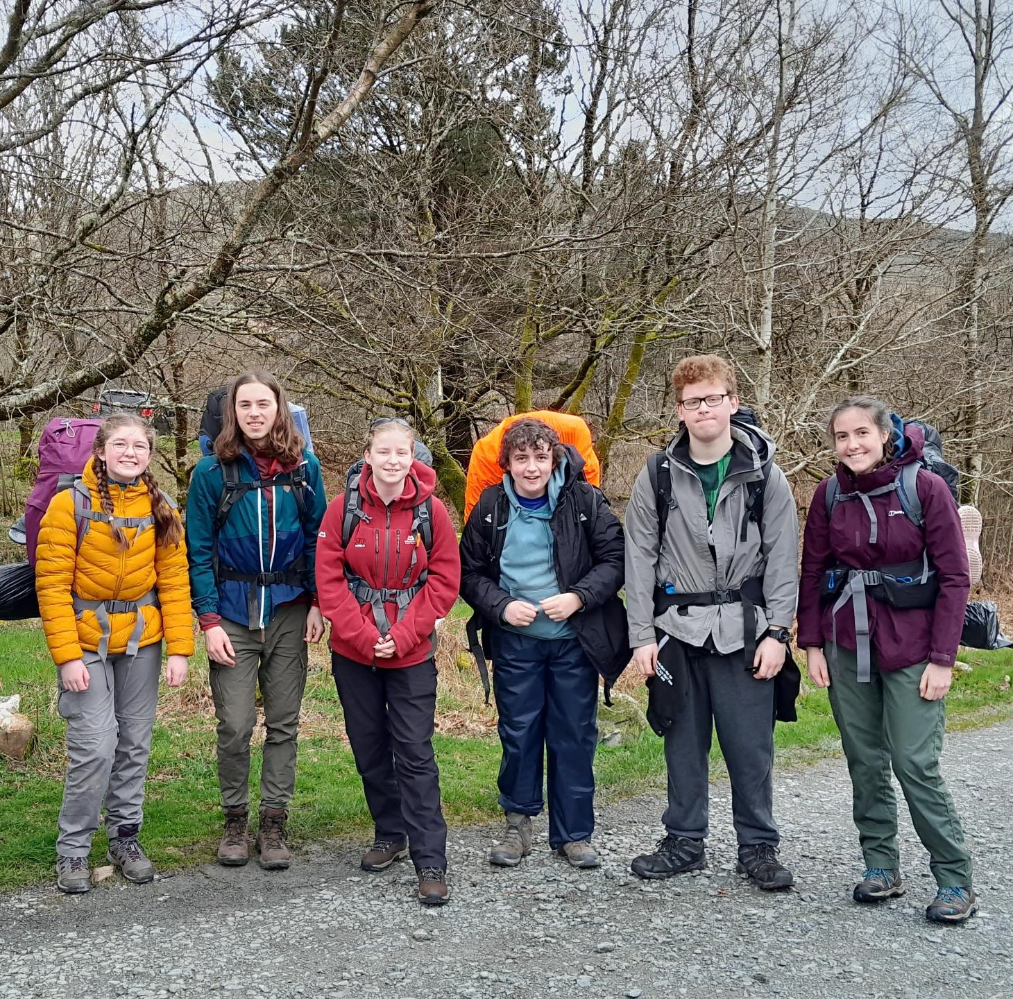 Six people with light skin are standing outdoors on a gravel path, posing for a group photo during a hiking trip. They are all dressed in outdoor gear, including jackets, hiking boots, and backpacks. From left to right: the first person is wearing a yellow jacket and gray pants with a large purple backpack; the second person has long brown hair, a teal jacket, and dark pants; the third person wears a red jacket, black pants, and smiles at the camera; the fourth person, standing slightly behind, is in a blue jacket and black pants with an orange backpack; the fifth person, wearing glasses, a gray jacket, and black pants, has hands in pockets; the sixth person on the right wears a burgundy jacket, green pants, and a gray backpack. Leafless trees and grassy hills surround them, suggesting a colder season or early spring.