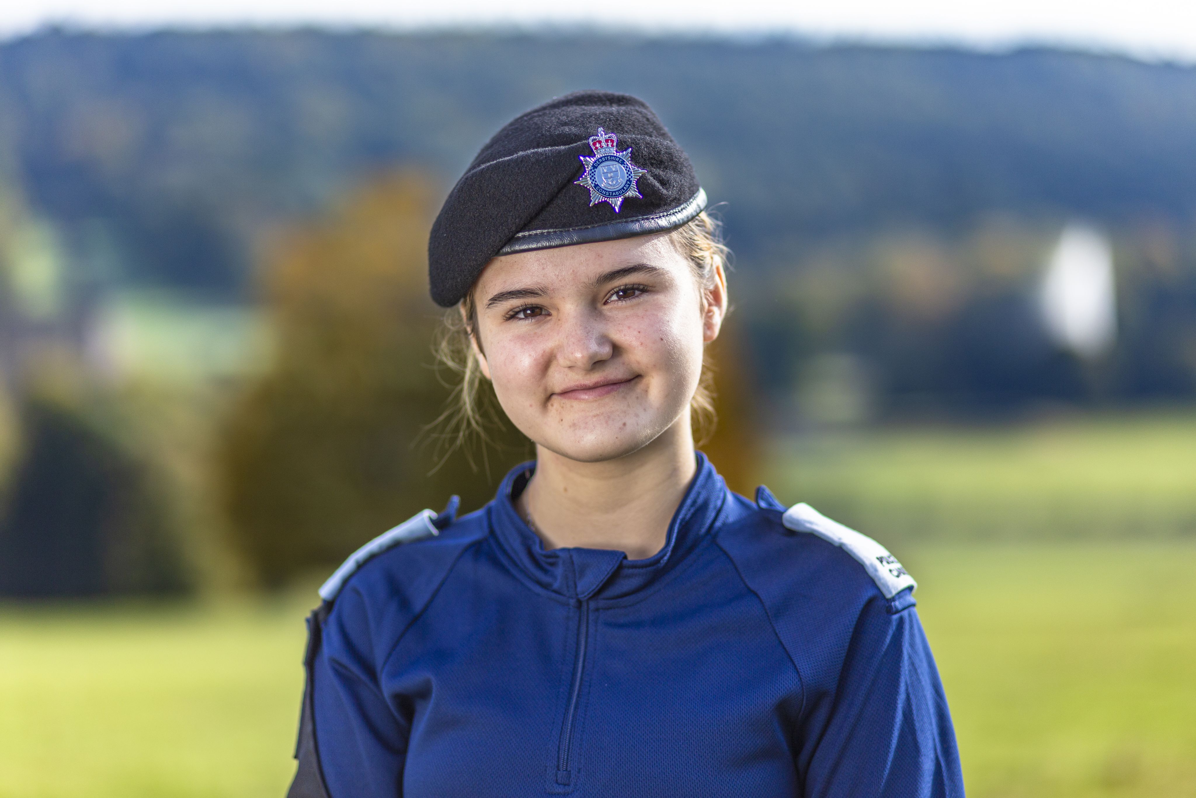 Amelie wearing a dark blue uniform and a black beret with a police insignia smiles at the camera. She stands outdoors with a blurred green landscape in the background, featuring trees and rolling hills. The lighting is bright, suggesting a sunny day.