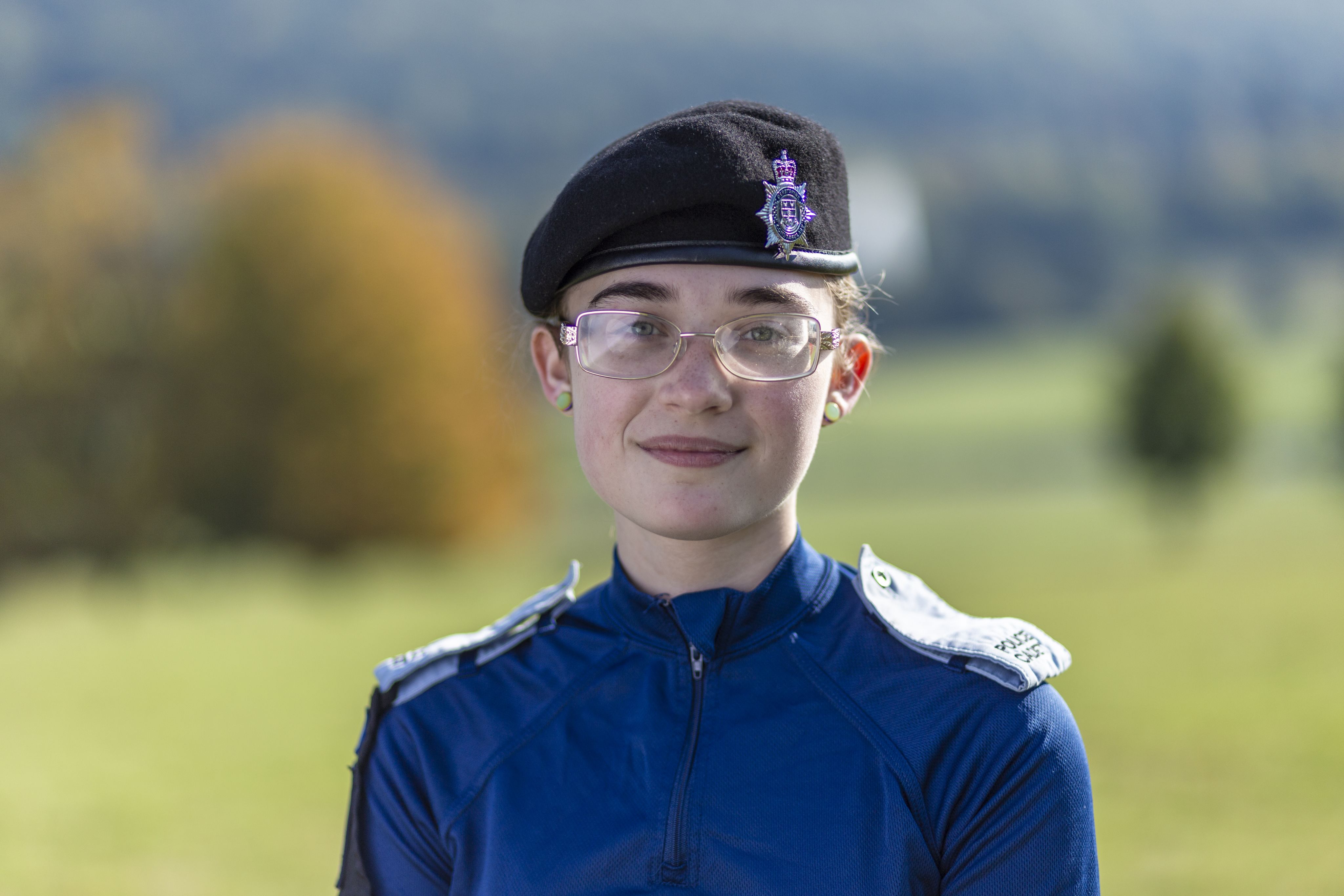 Teri wearing a dark blue uniform and a black beret with a police insignia smiles at the camera. She stands outdoors with a blurred green landscape in the background, featuring trees and rolling hills. The lighting is bright, suggesting a sunny day.