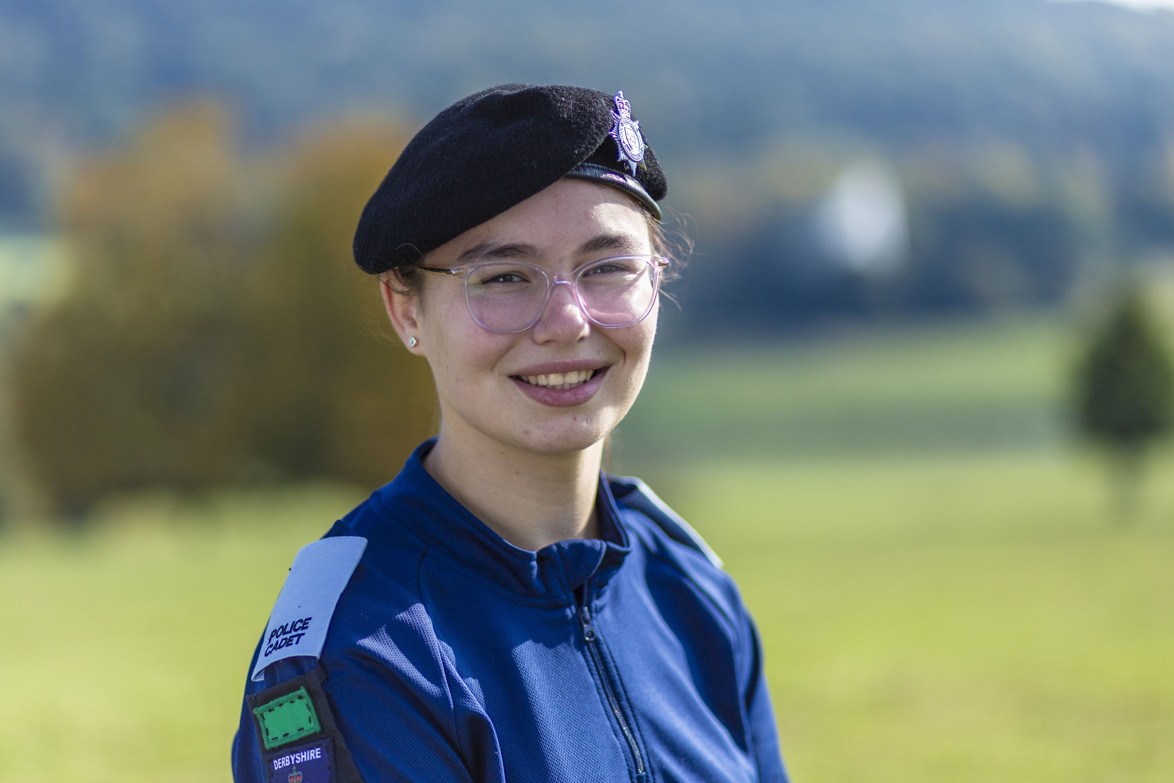Bethan wearing a dark blue uniform and a black beret with a police insignia smiles at the camera. She stands outdoors with a blurred green landscape in the background, featuring trees and rolling hills. The lighting is bright, suggesting a sunny day.