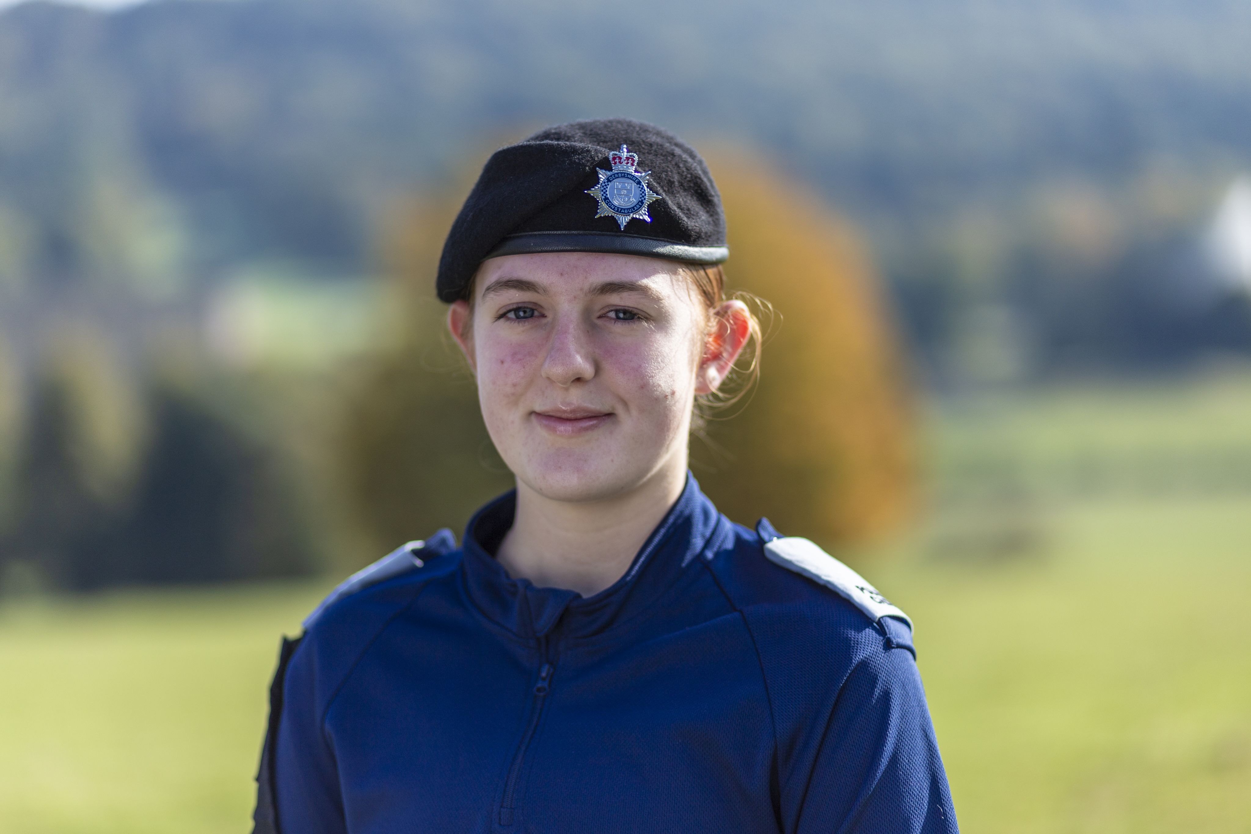 Clarissa wearing a dark blue uniform and a black beret with a police insignia smiles at the camera. She stands outdoors with a blurred green landscape in the background, featuring trees and rolling hills. The lighting is bright, suggesting a sunny day.