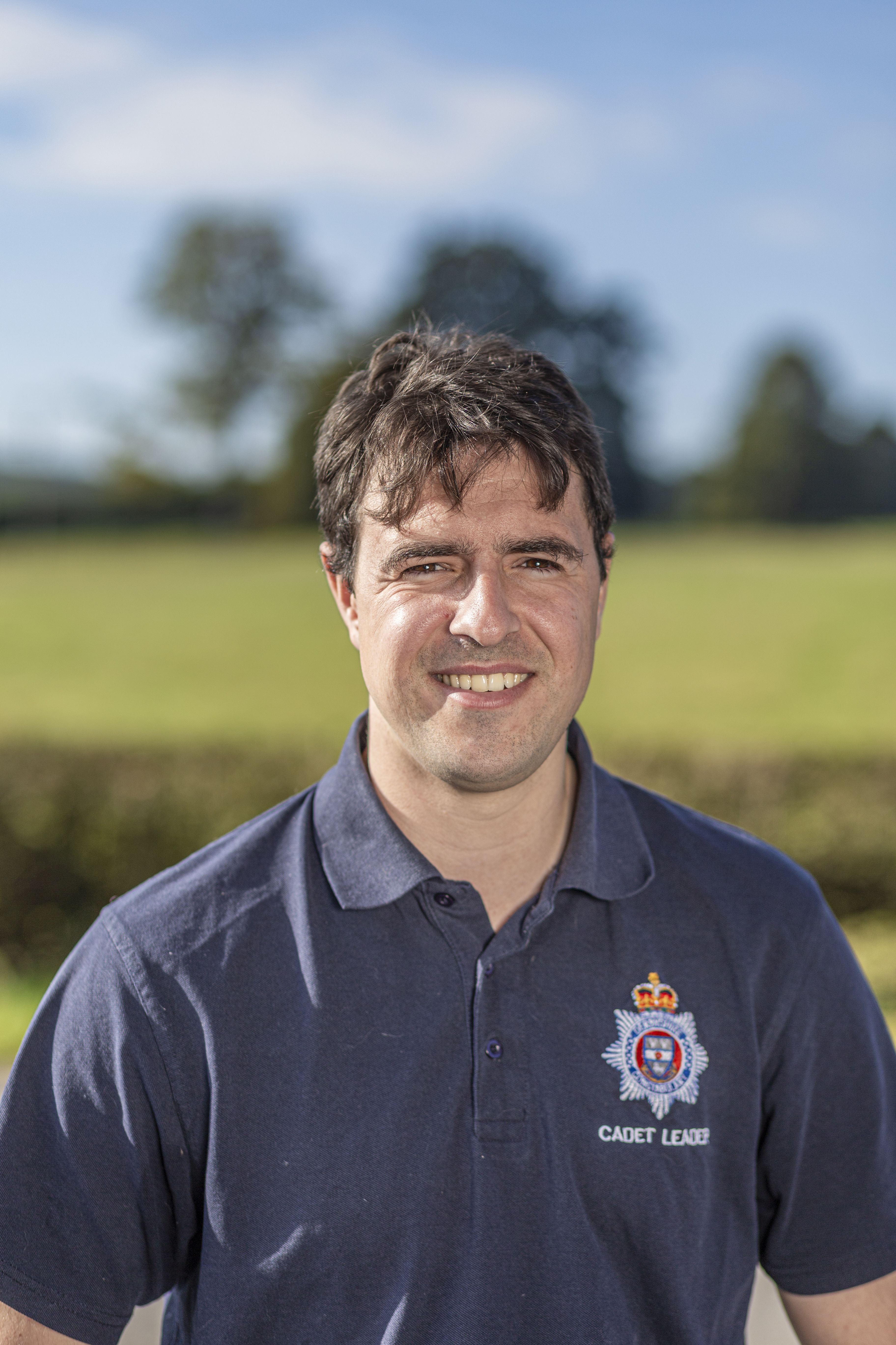 Dom stands outdoors, wearing a navy blue polo shirt with a badge that reads "CADET LEADER." The background features a green field and a clear blue sky.
