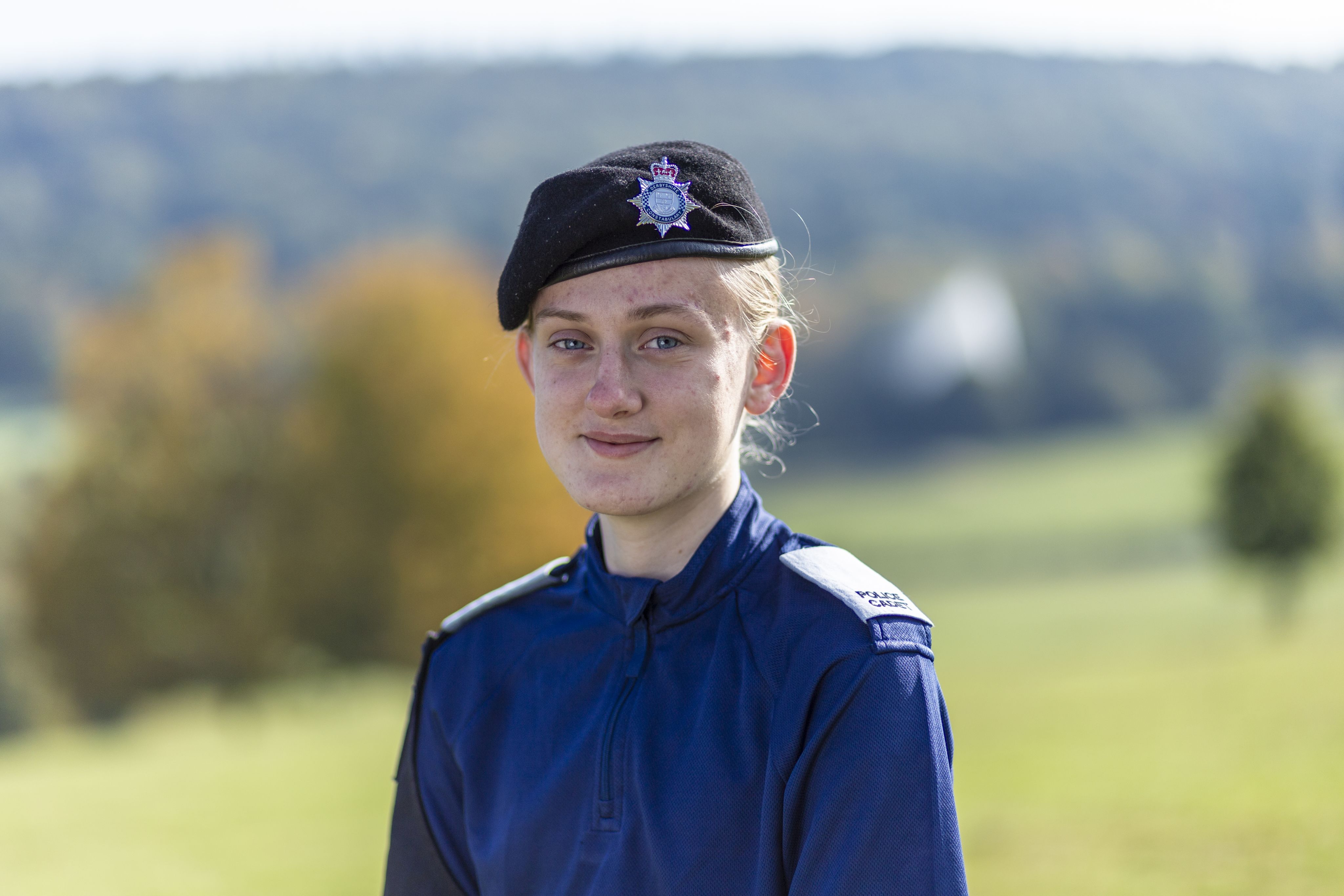 Emilia wearing a dark blue uniform and a black beret with a police insignia smiles at the camera. She stands outdoors with a blurred green landscape in the background, featuring trees and rolling hills. The lighting is bright, suggesting a sunny day.