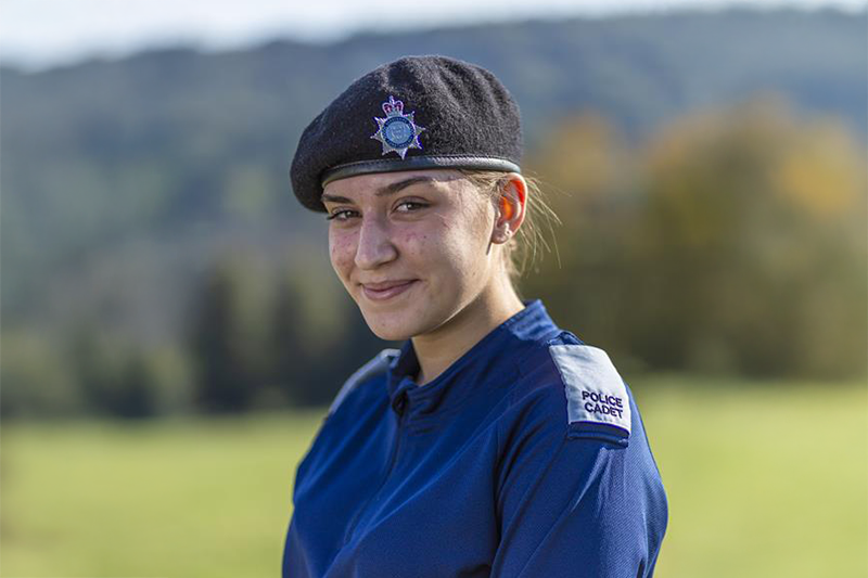 Rosie wearing a dark blue uniform and a black beret with a police insignia smiles at the camera. She stands outdoors with a blurred green landscape in the background, featuring trees and rolling hills. The lighting is bright, suggesting a sunny day.