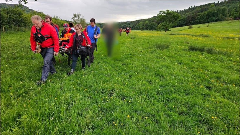 A group of people, dressed in outdoor clothing, are carrying a stretcher through a grassy field. The individuals at the front and sides are wearing red jackets, while others in the background are visible walking behind. The lush green field is dotted with small yellow flowers, and there are trees and rolling hills in the background under an overcast sky. One individual is blurred on the stretcher, indicating a rescue or assistance scenario. The group appears focused and coordinated as they move through the open landscape.