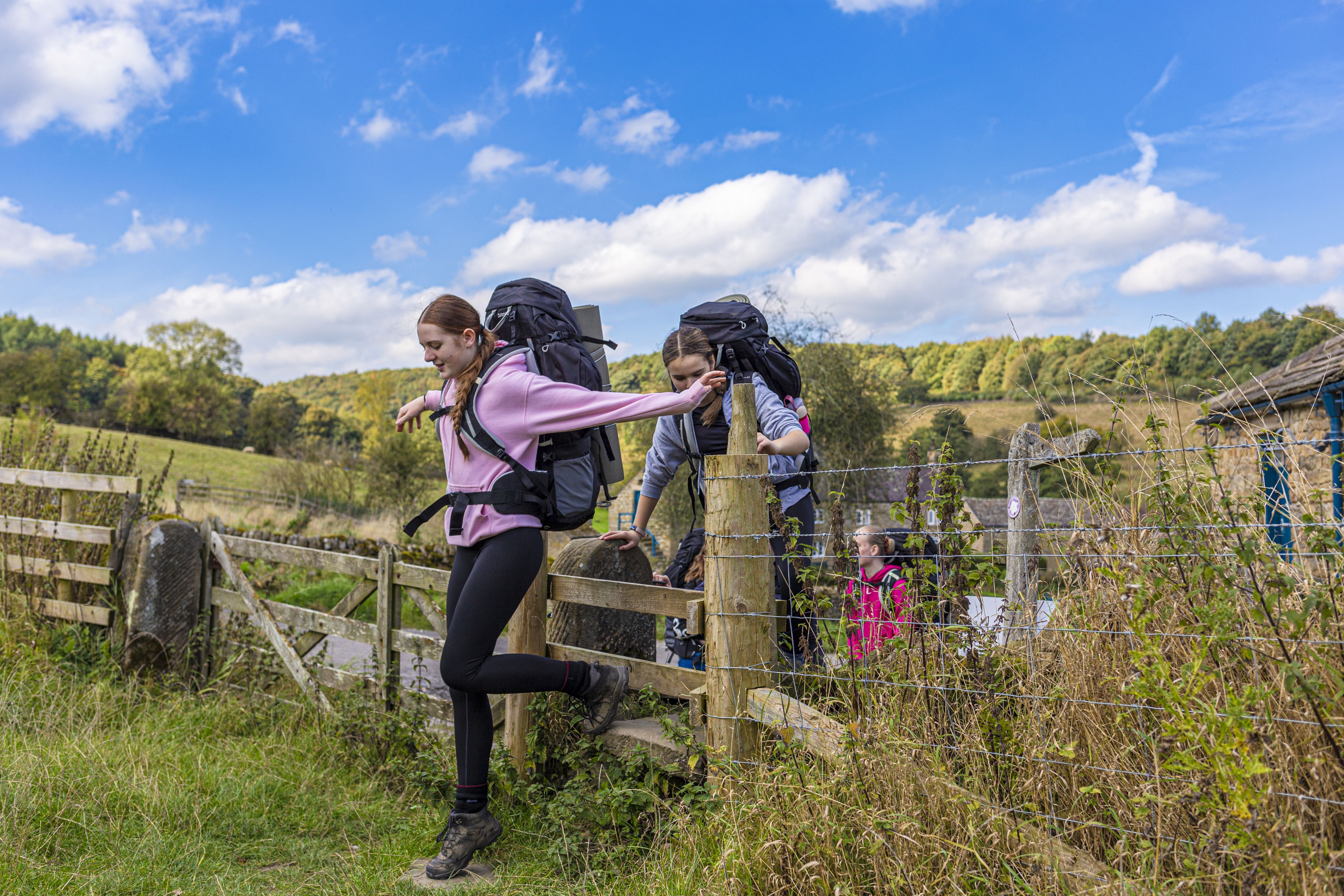 A young woman in a pink sweater and black leggings is climbing over a wooden fence while carrying a large backpack. In the background, two other individuals, also with backpacks, are navigating the fence. The scene is set in a lush, green landscape with trees and a blue sky dotted with clouds.