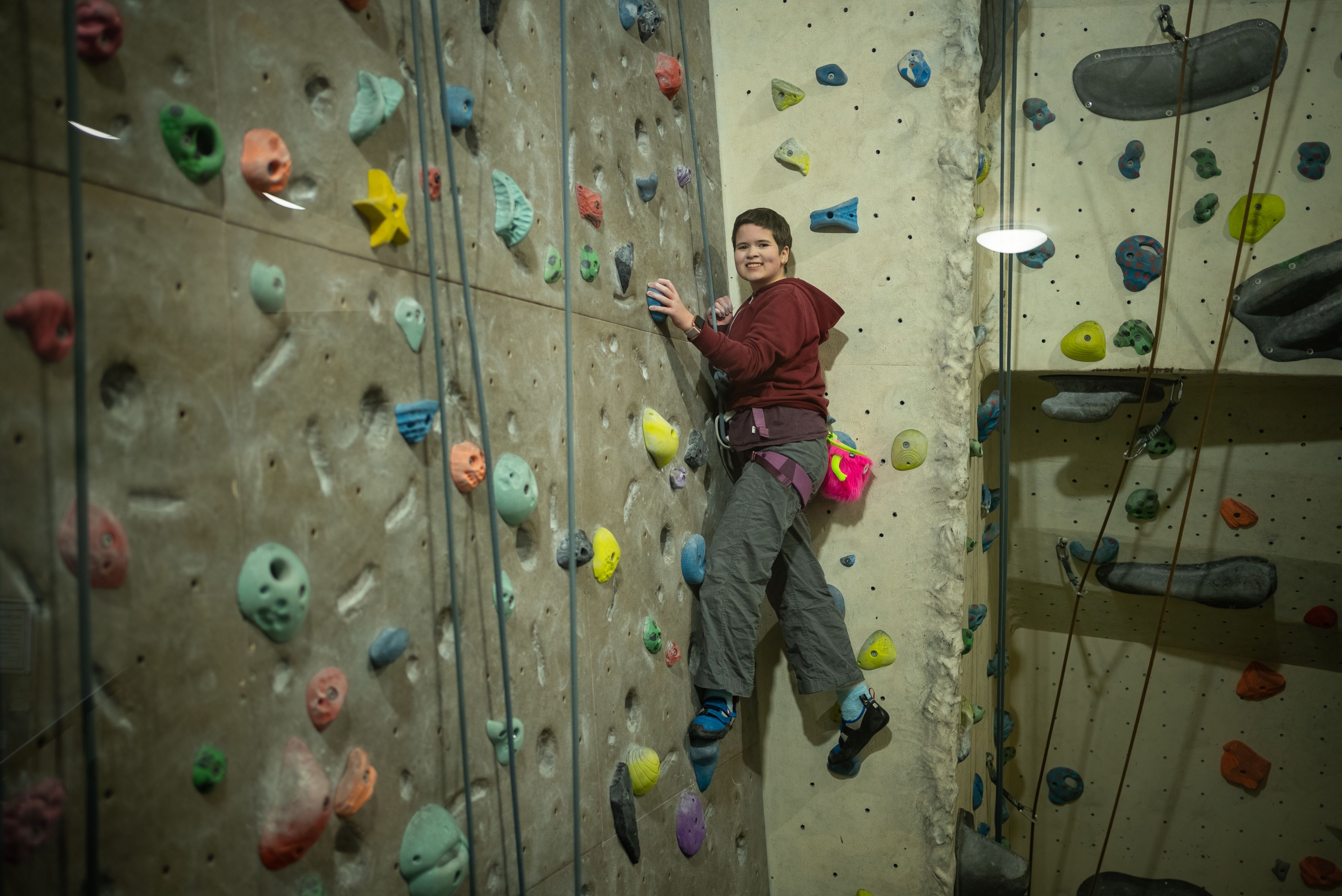 Rosa climbing a colorful indoor rock climbing wall. She is wearing a maroon hoodie and gray climbing pants, with climbing shoes and a harness. The wall is adorned with various holds in different shapes and colors, and Rosa is positioned mid-climb, smiling confidently.