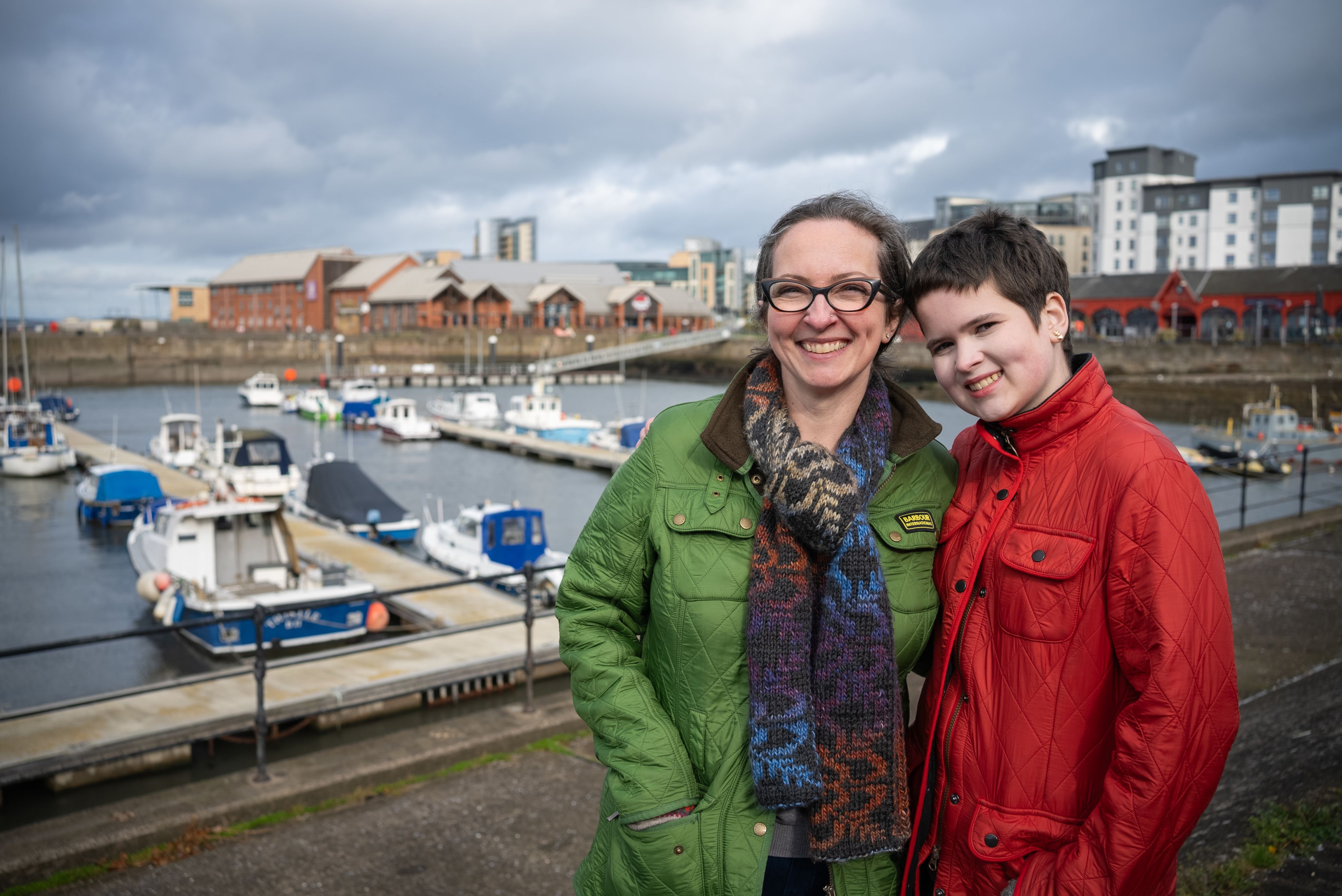 Rachel and Rosa stand smiling together at a harbor. Rachel is wearing a green jacket and glasses, while Rosa is dressed in a bright red jacket. In the background, there are several boats docked in the water and buildings lining the shore under a cloudy sky.