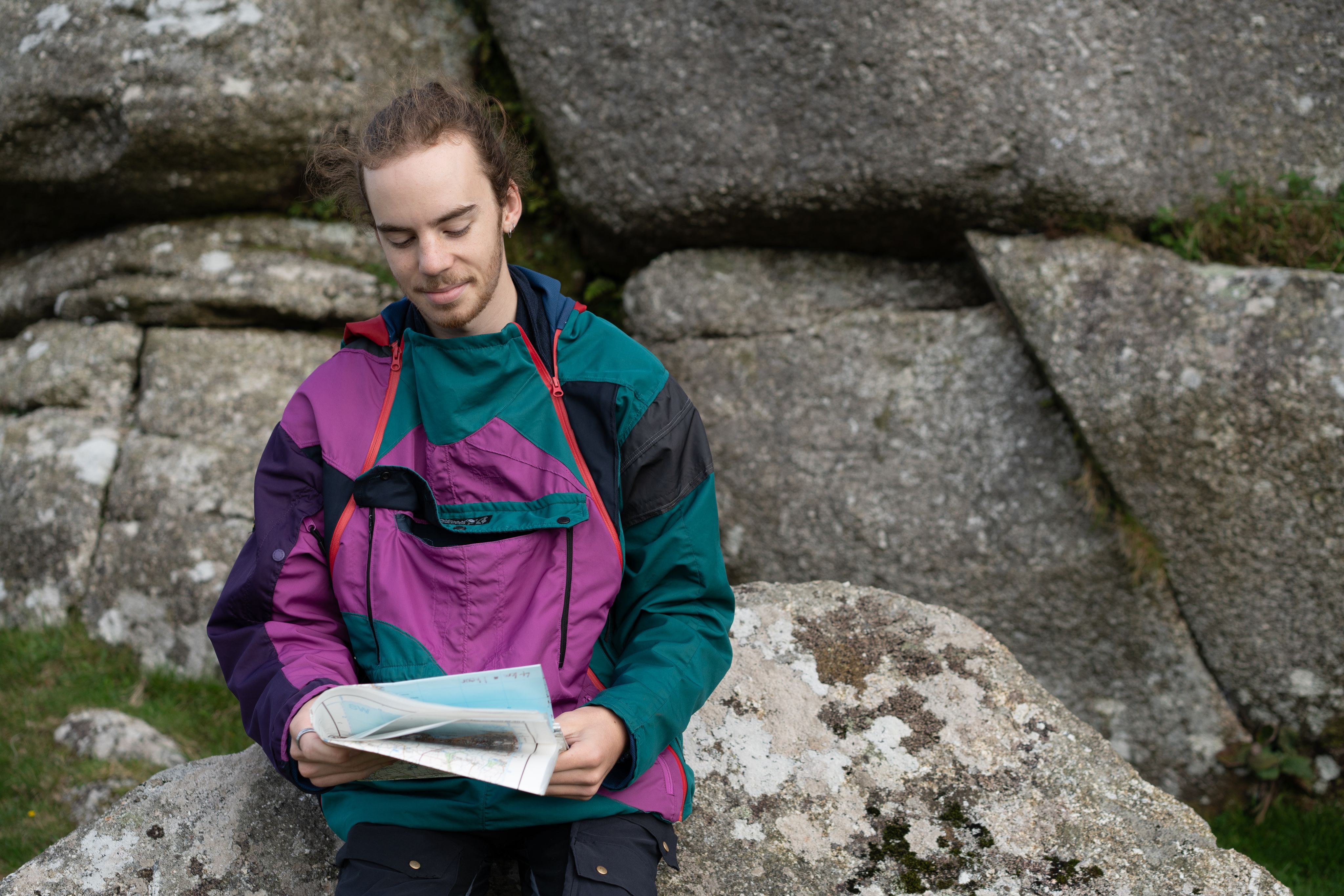 Josh sits on a large rock, wearing a colorful jacket with shades of purple, teal, and black. He is reading a book or map, looking relaxed and content, with a backdrop of large, textured boulders. The setting appears to be outdoors, possibly in a natural or rugged environment.