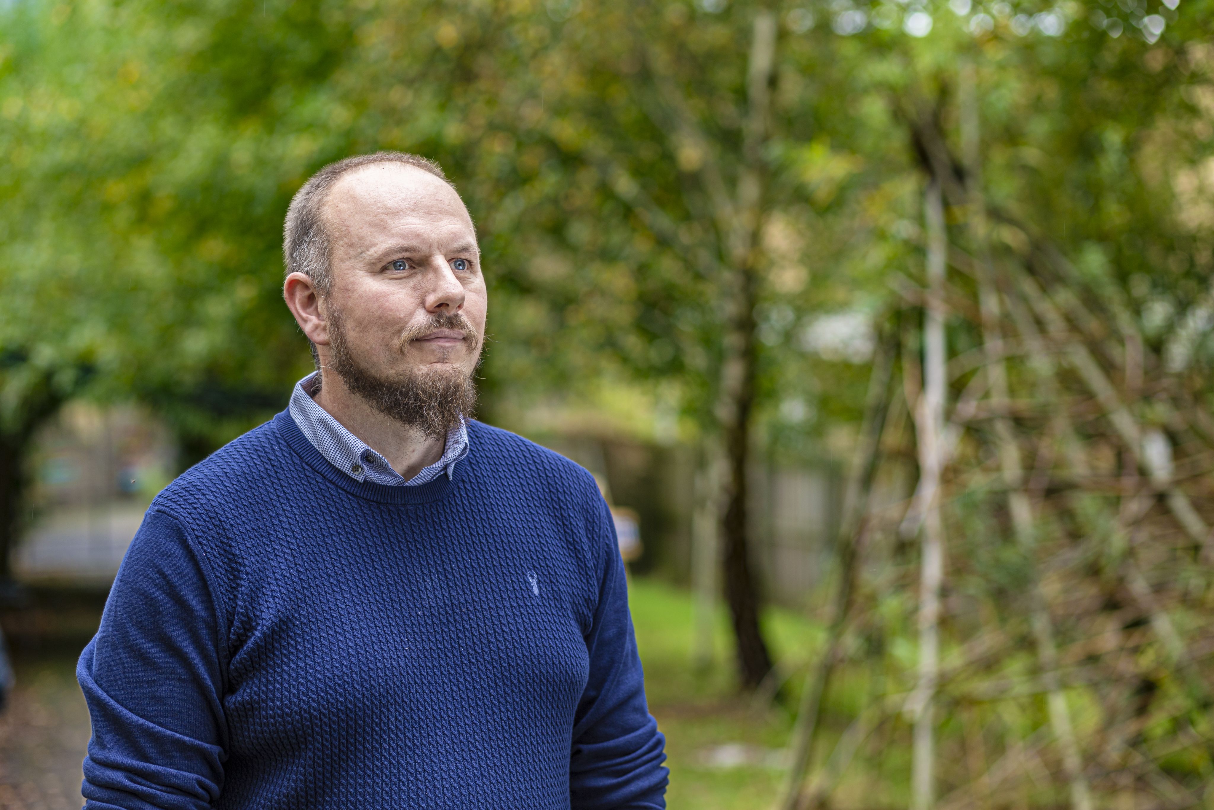 Danny stands outdoors, looking thoughtfully into the distance. He is wearing a blue sweater and a collared shirt. The background features greenery and trees, creating a natural setting.
