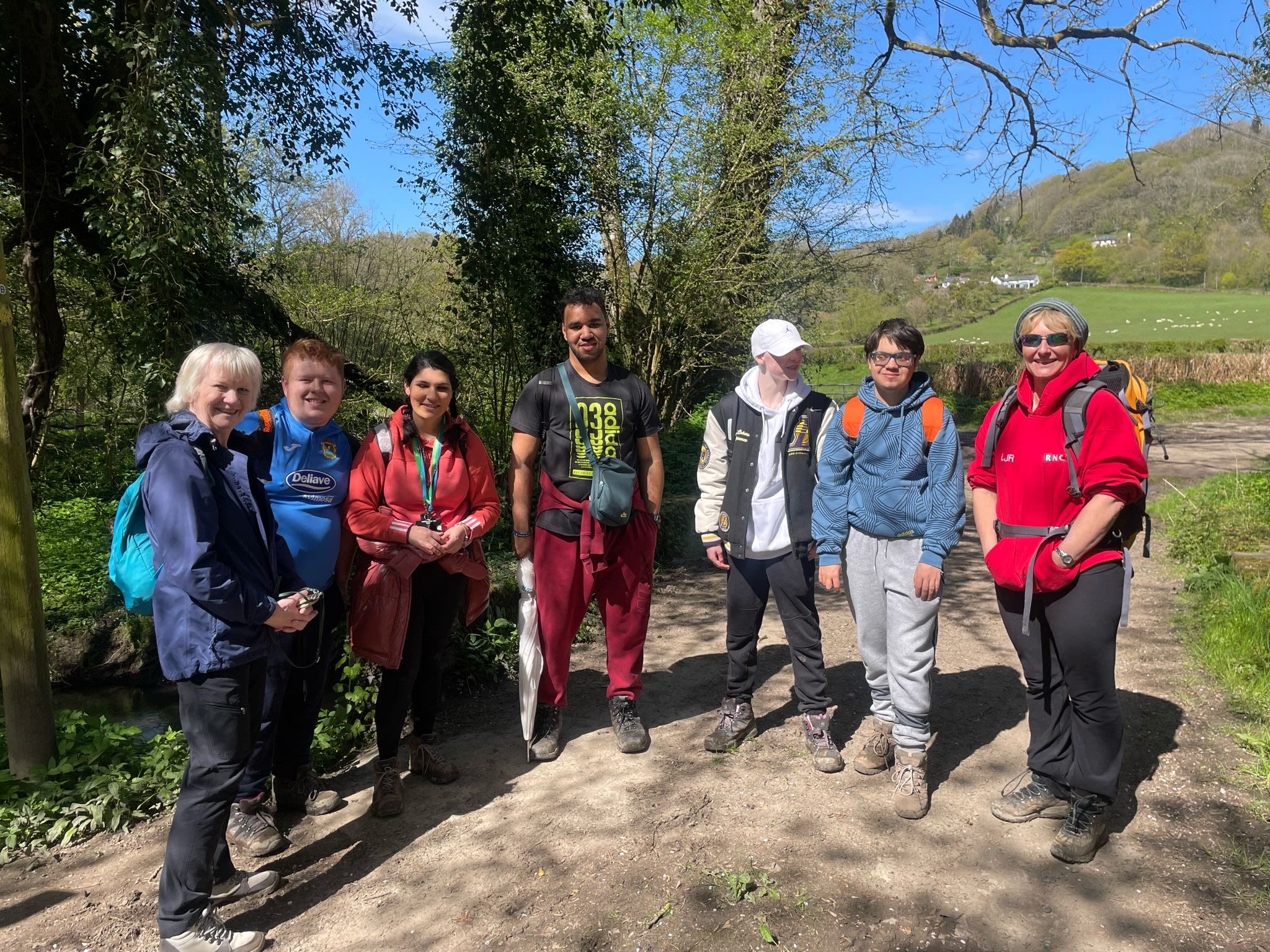 A group of seven people stands on a dirt path surrounded by green trees and hills on a sunny day. The group consists of individuals of varying ages, genders, and ethnicities wearing outdoor clothing, including jackets, hoodies, and walking shoes. Two people wear backpacks, and one is holding a walking pole. They are all smiling or looking relaxed, standing close together, as they pose for a group photo during a hike or outdoor activity. The background features a countryside landscape with rolling hills, trees, and clear blue skies.
