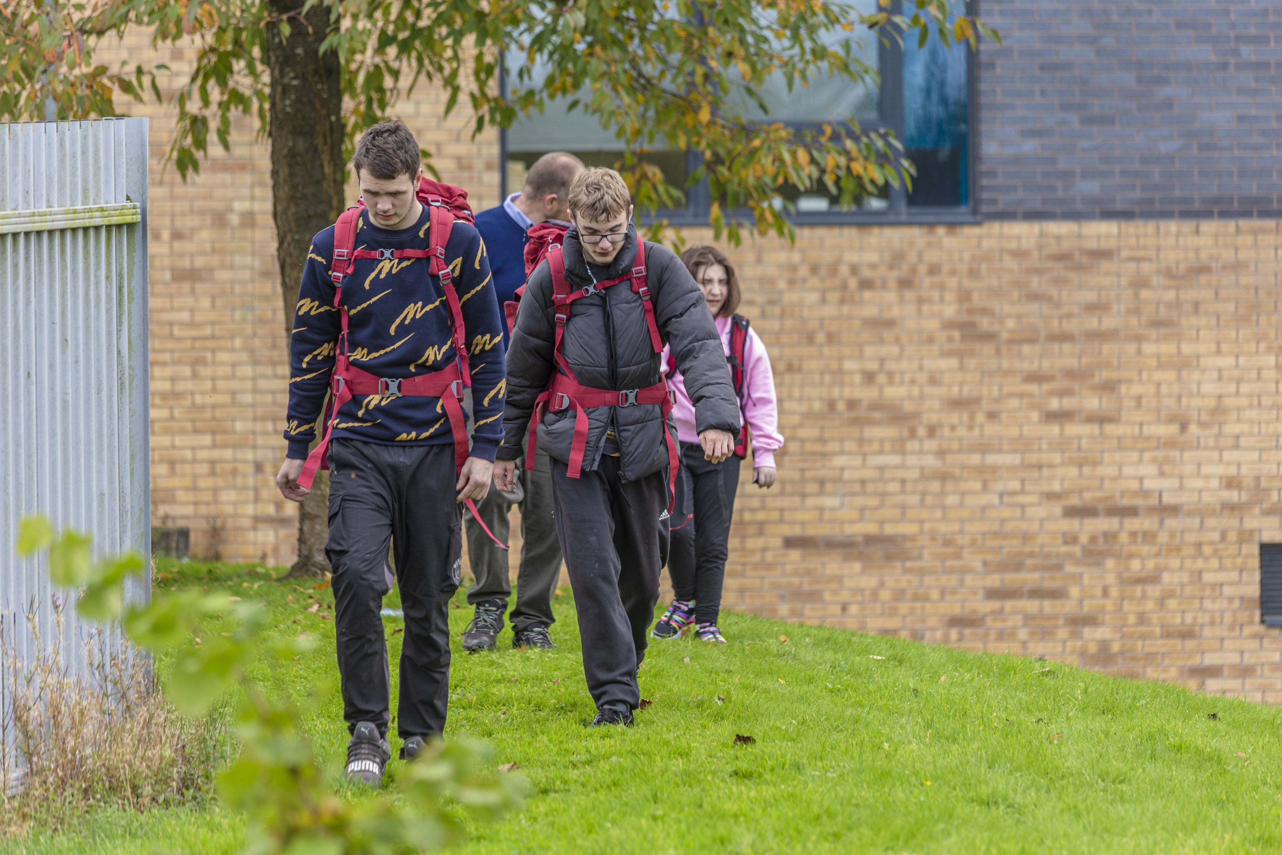 A group of four individuals, wearing backpacks and outdoor clothing, walks along a grassy area near a building with a brick facade. The first two individuals are focused on the ground as they walk, while the others follow closely behind. Trees and a fence are visible in the background.