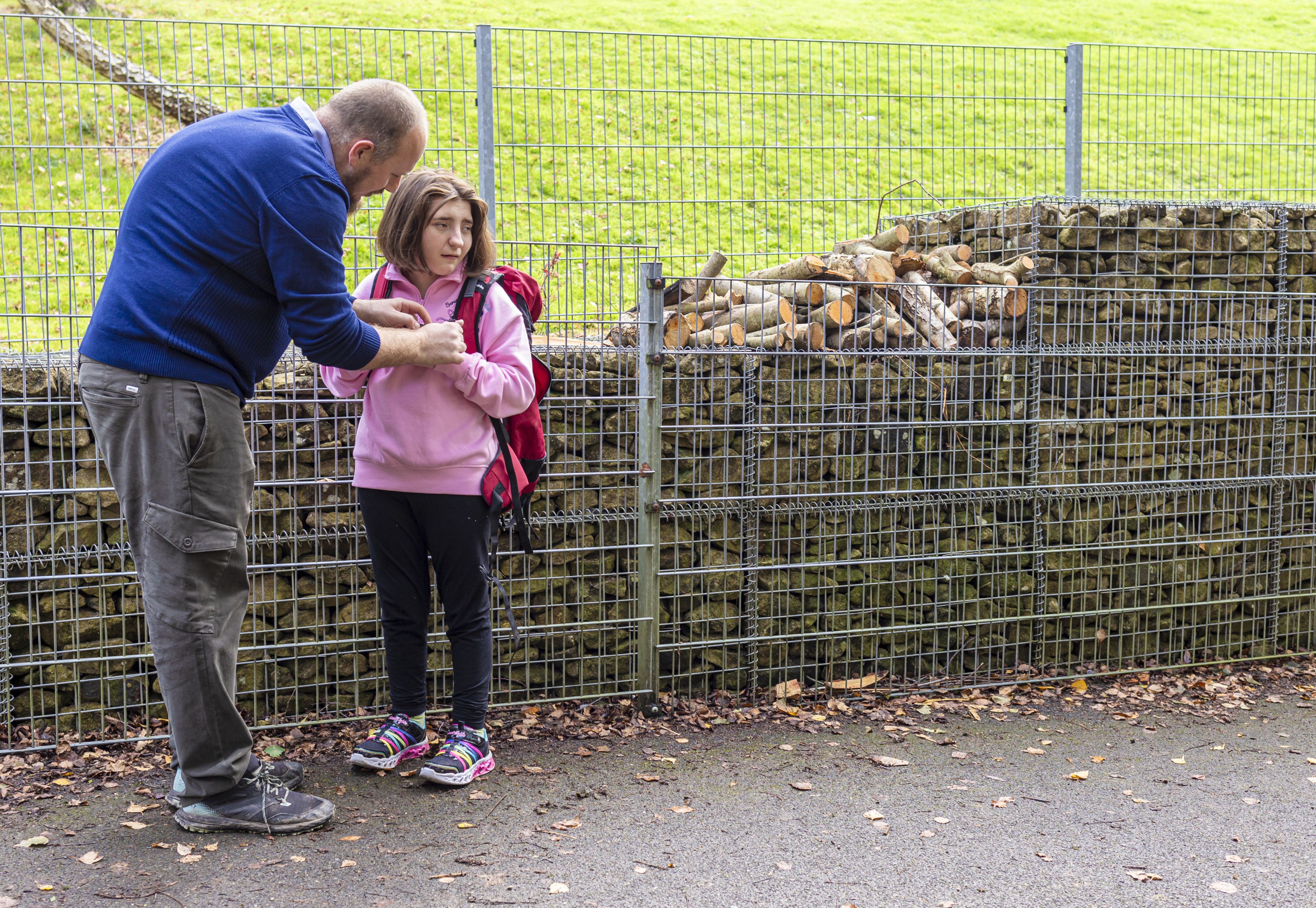 Danny is adjusting a backpack on Ruby standing beside a stone wall. The girl looks slightly apprehensive, while the man appears focused on helping her. The background features a grassy area and a fence.