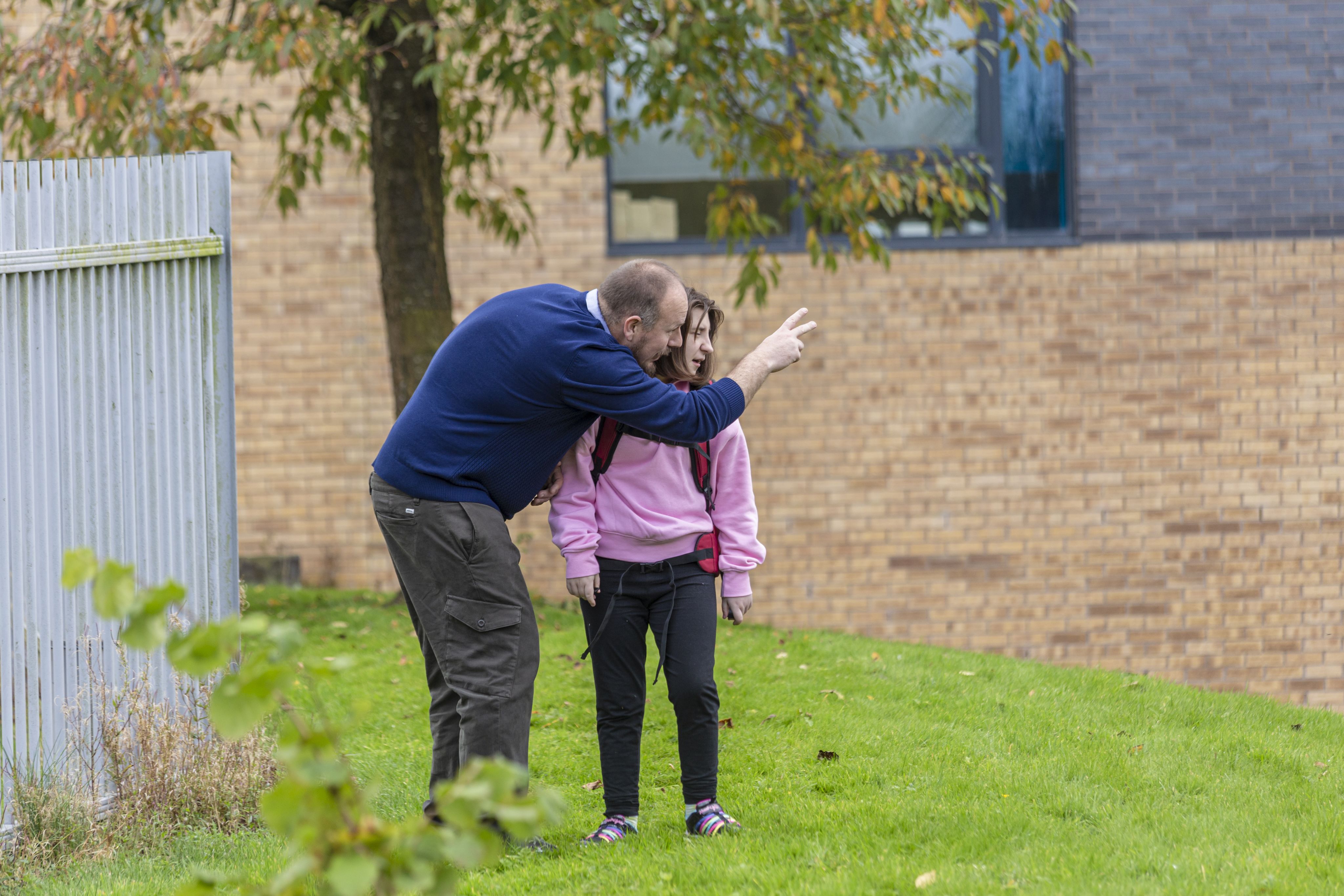 Danny leans down to speak to Ruby, pointing in a direction while she looks up at him. They are standing on a grassy area near a building with a brick facade. The girl is wearing a pink hoodie and has a backpack, while the man is dressed in a blue sweater and brown pants. Trees with green leaves are visible in the background.