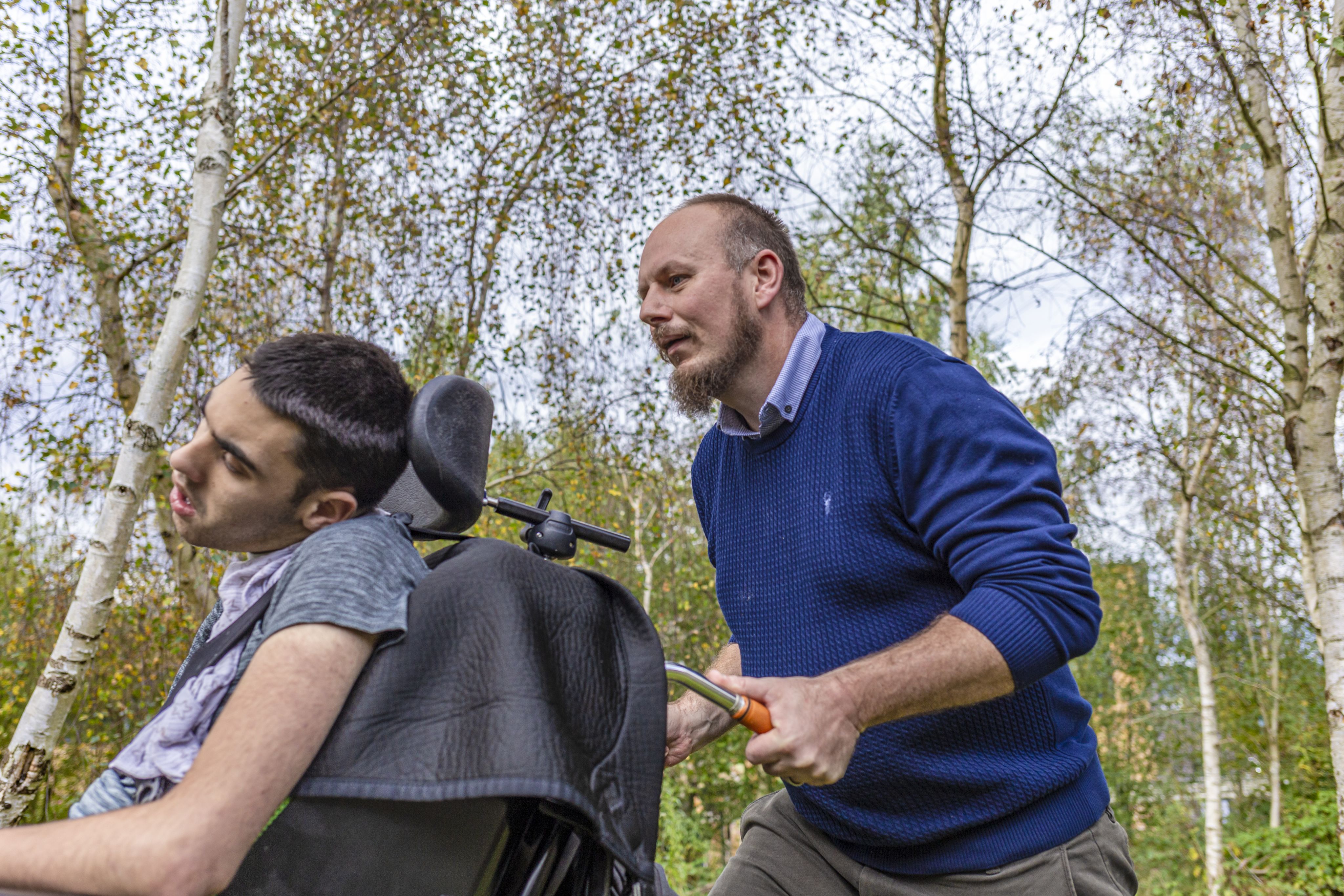 Danny pushing a wheelchair through a park filled with trees. The person in the wheelchair, a young man, appears to be looking to the side. The background features green foliage and a clear sky, suggesting a pleasant outdoor setting.