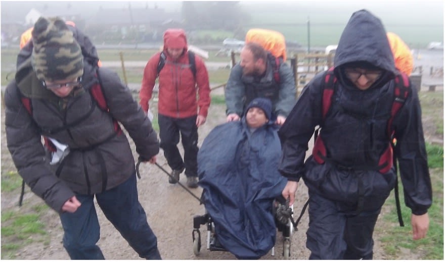 A group of five individuals is hiking on a muddy trail in foggy weather. Two people at the front are pulling a specialized wheelchair with a person seated, covered by a waterproof poncho. Behind them, two others are helping push the wheelchair, and all are wearing rain gear and backpacks with waterproof covers. The foggy landscape and rural setting are visible in the background. The group appears focused and working together to navigate the terrain.