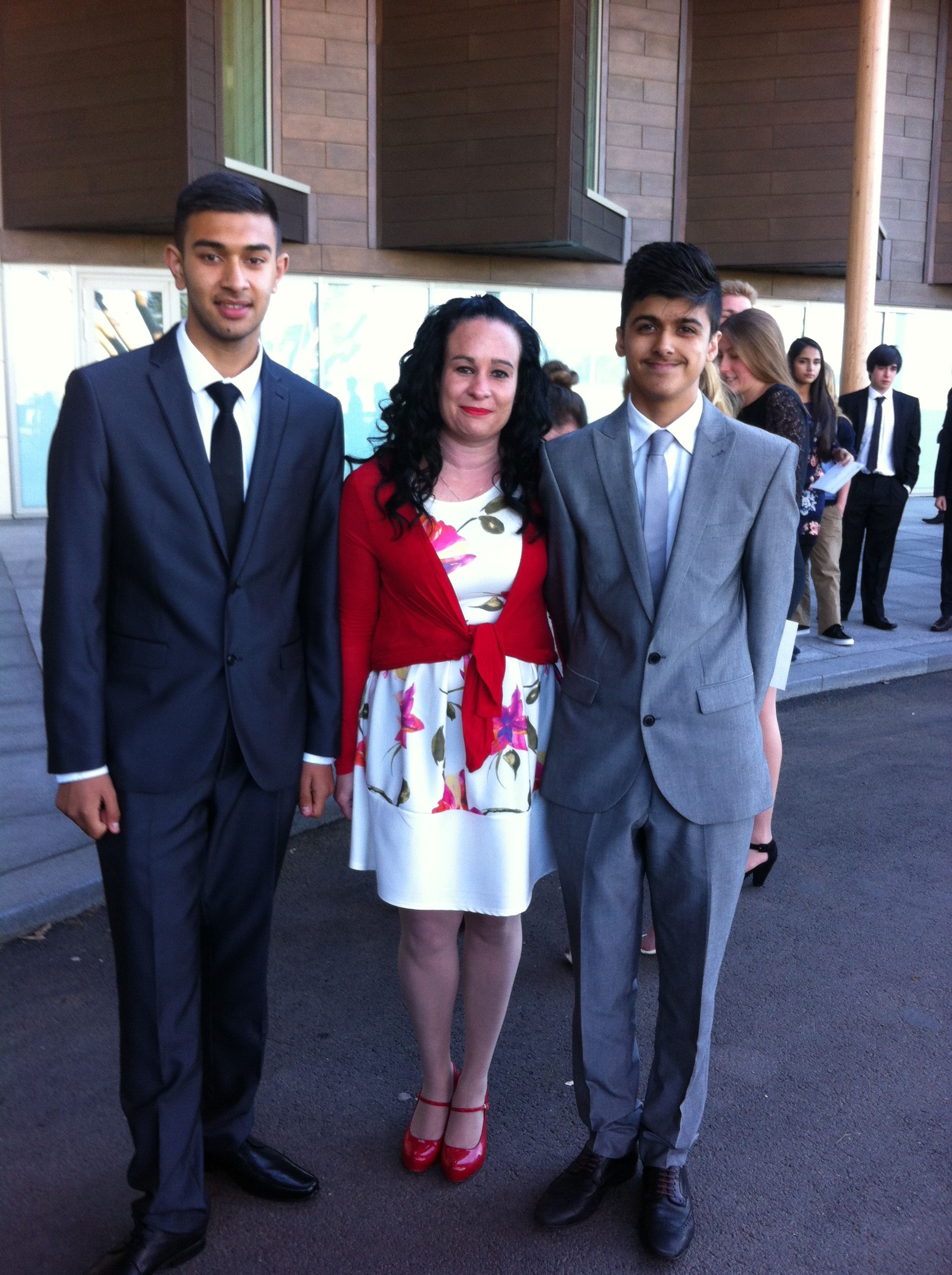 Two young men and a woman are posing outside a modern building with large glass windows and wooden panels. The young men are wearing formal suits—one in dark gray and the other in light gray—paired with white shirts and ties. The woman standing between them is wearing a floral white dress with a red cardigan. All three are smiling at the camera. Other people dressed in formal attire can be seen in the background, suggesting a formal event, such as a graduation or awards ceremony.