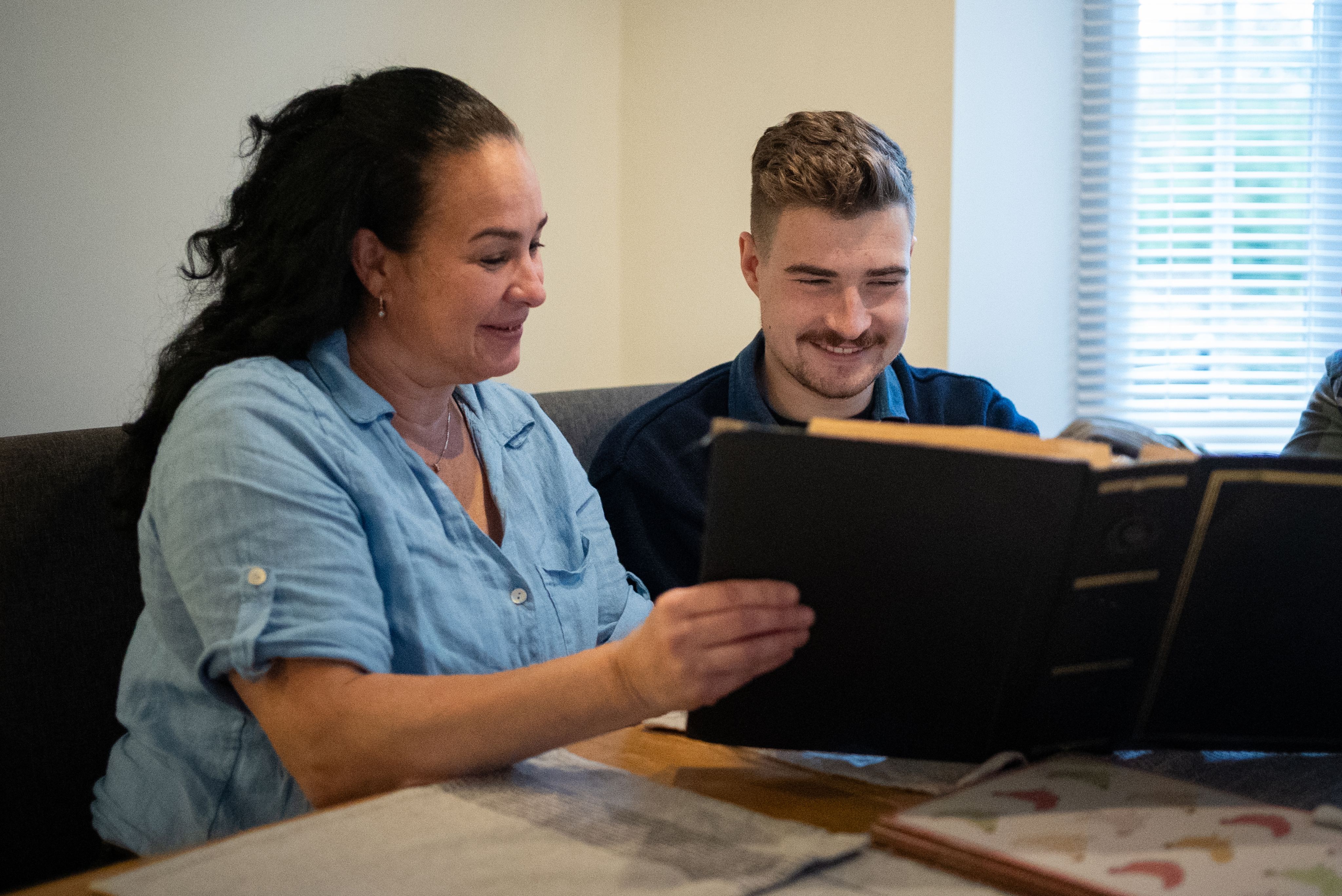 Lisa and her son are sitting at a table, looking at a large photo album together. Lisa, with long dark hair, is smiling as she points to a page in the album, while the man, with short light hair and a mustache, is also smiling and engaged in the moment. The setting appears to be a cozy indoor space with natural light coming through a window.