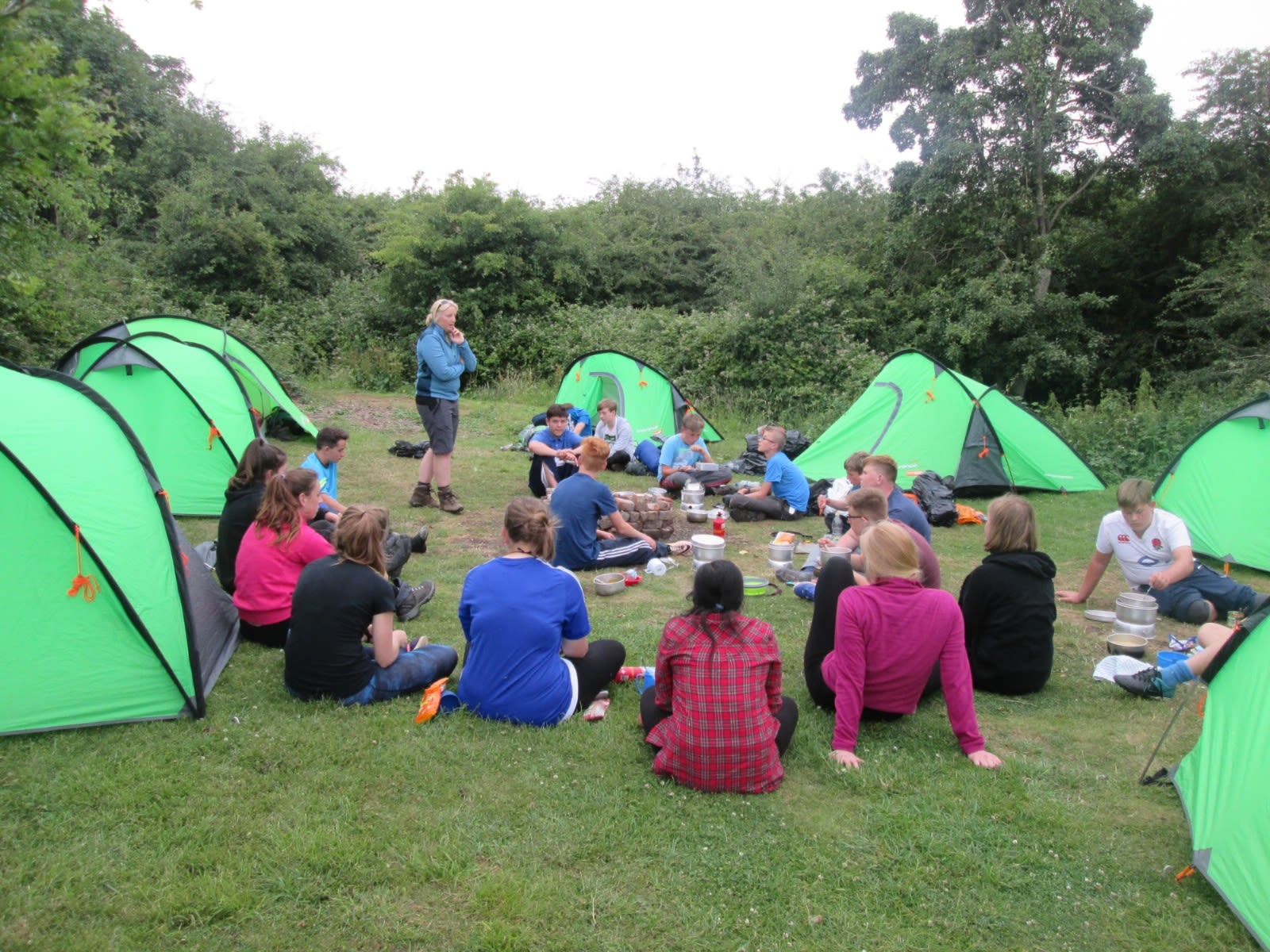A group of young people and an adult supervisor are gathered outdoors in a grassy area surrounded by bushes and trees. Several green tents are set up in the background, suggesting a camping trip. The young people are sitting in a circle on the grass, engaged in a group activity or discussion, with some cooking equipment and food items in the center. The adult supervisor, wearing a blue jacket and boots, is standing to the left, addressing the group. The scene reflects a Duke of Edinburgh's Award experience.