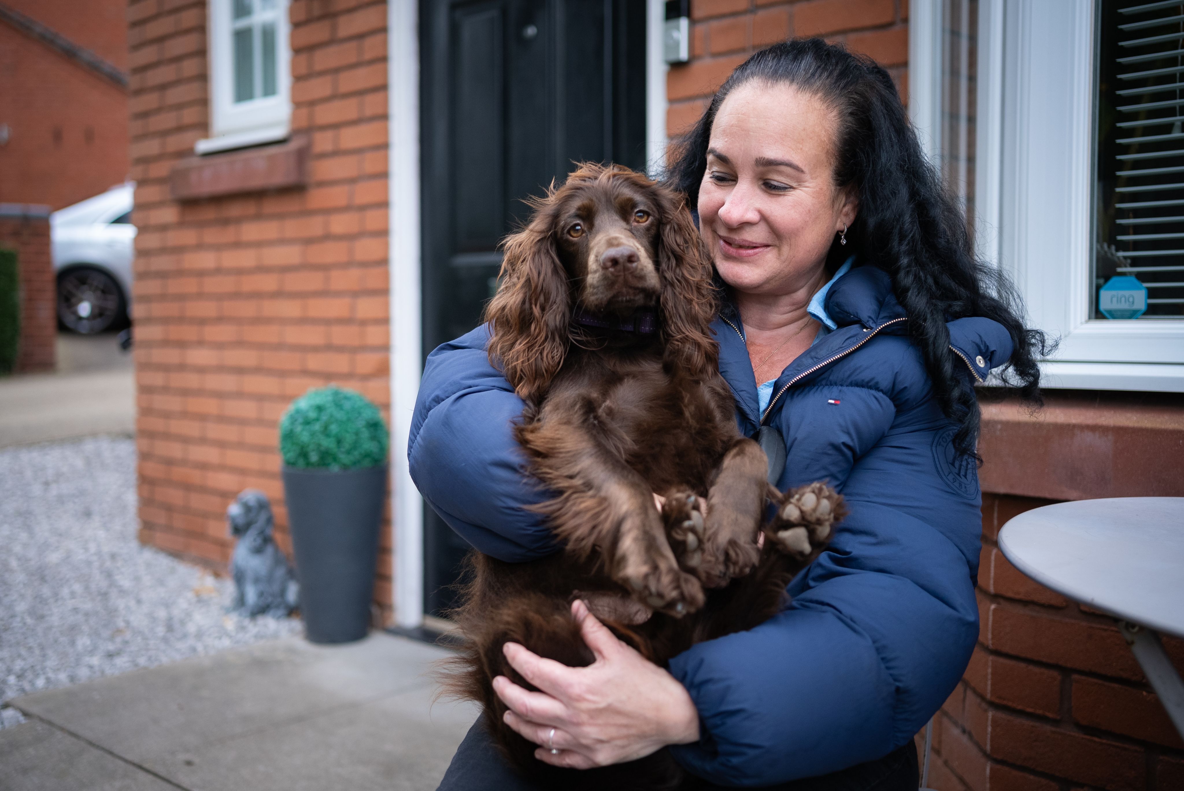 Lisa with long, dark hair is holding a brown spaniel dog in her arms, smiling at the camera. They are standing outside a brick house, with a potted plant nearby and a glimpse of a car in the background. The woman is wearing a blue puffer jacket.