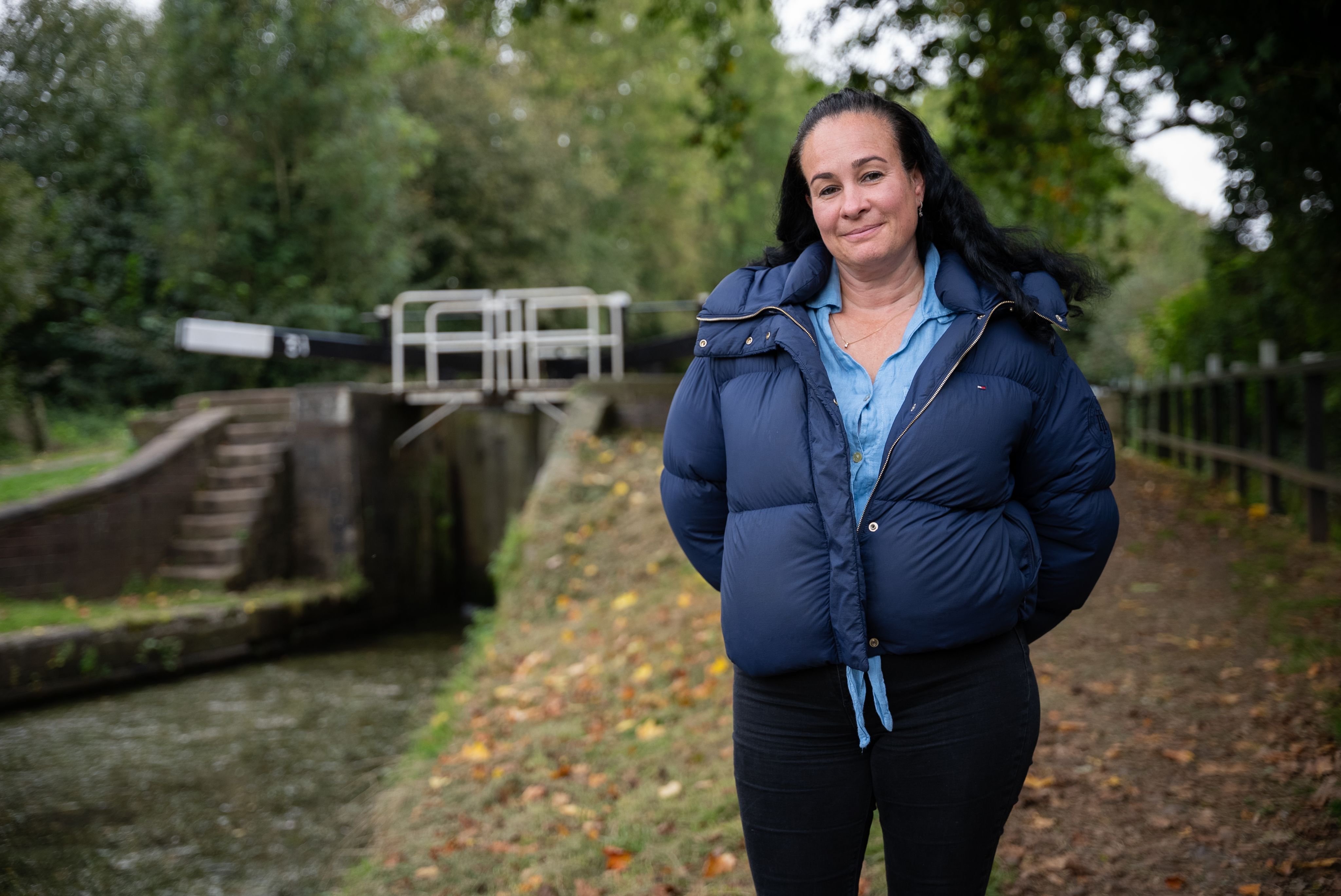 Lisa is standing outdoors near a canal lock. She is wearing a blue puffer jacket over a light blue shirt and is smiling gently. The background features greenery and a canal with a lock gate visible.