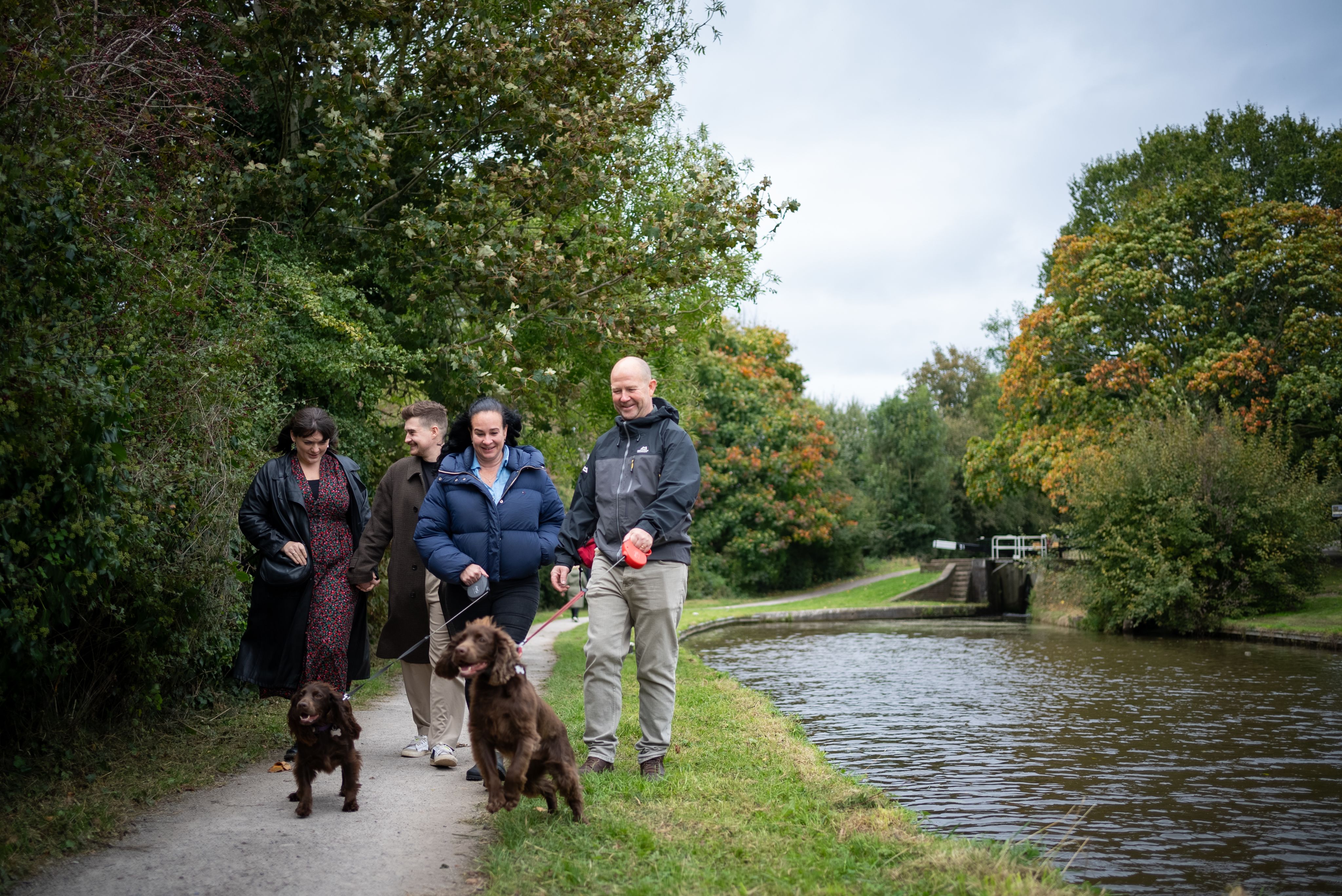 A group of four people walking along a canal path with two brown dogs. The scene features lush greenery and trees with autumn foliage. The individuals are casually dressed, and the atmosphere appears relaxed and friendly.